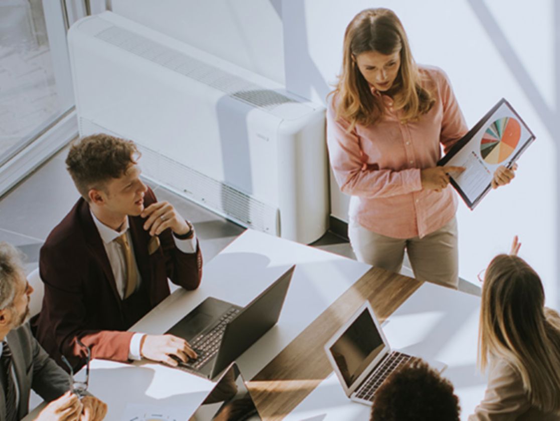 In a room, there are people sitting at a table and a woman standing showing them a pie chart.