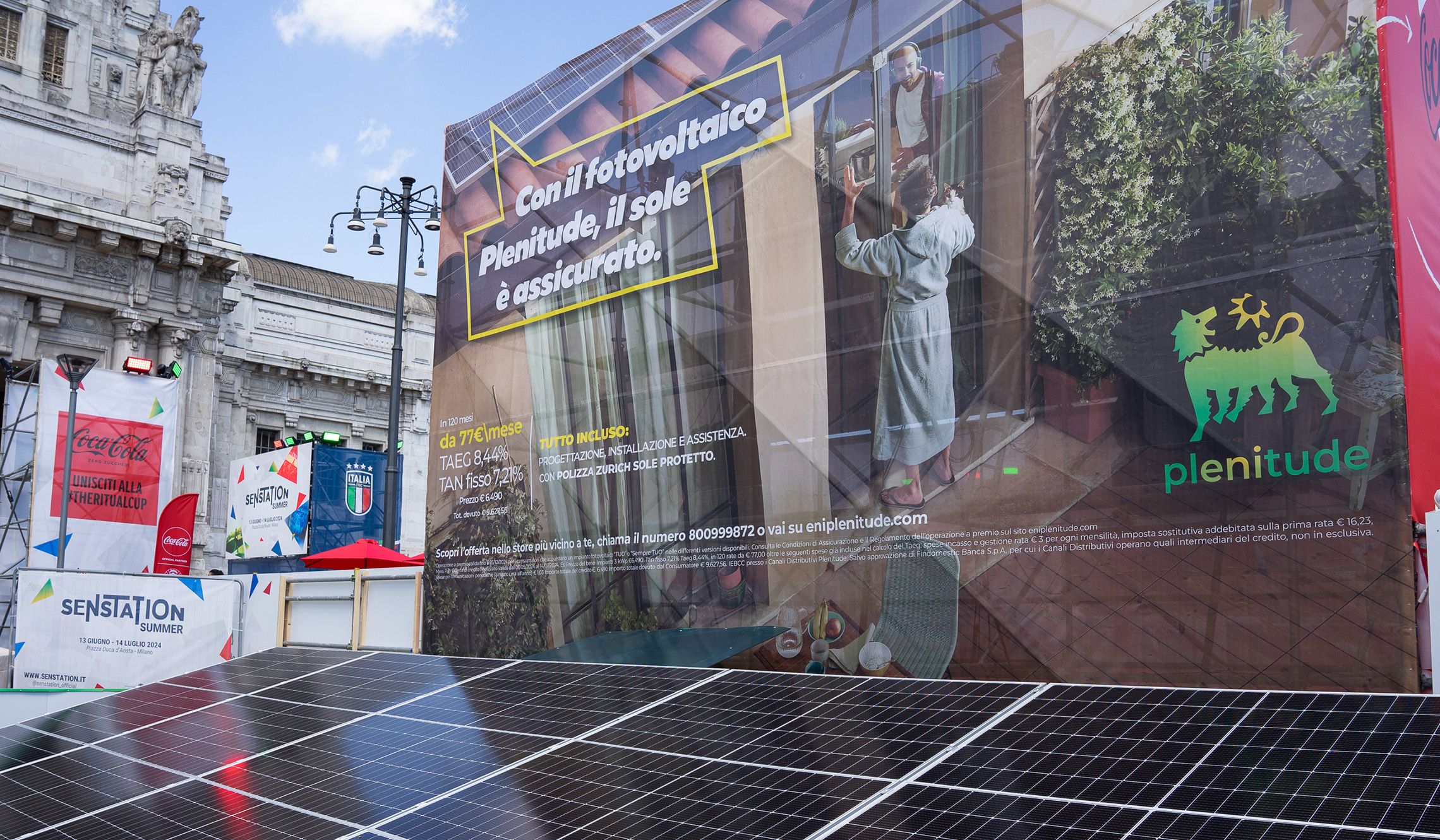 In primo piano ci sono dei pannelli solari e un cartellone con un’immagine che raffigura due ragazzi, il primo è dentro casa e il secondo è fuori dalla porta finestra in accappatoio, c’è anche il logo di Plenitude e una scritta che dice Con il fotovoltaico Plenitude, il sole è assicurato. Sullo sfondo, la stazione Centrale di Milano.  