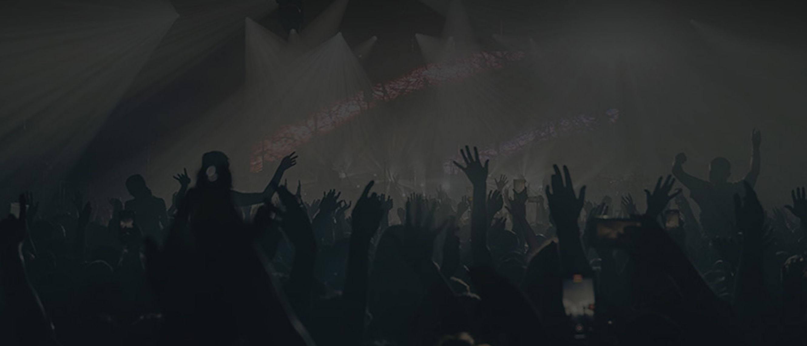 In an enclosed space there is a crowd with their hands raised and a stage with coloured lights in the background.