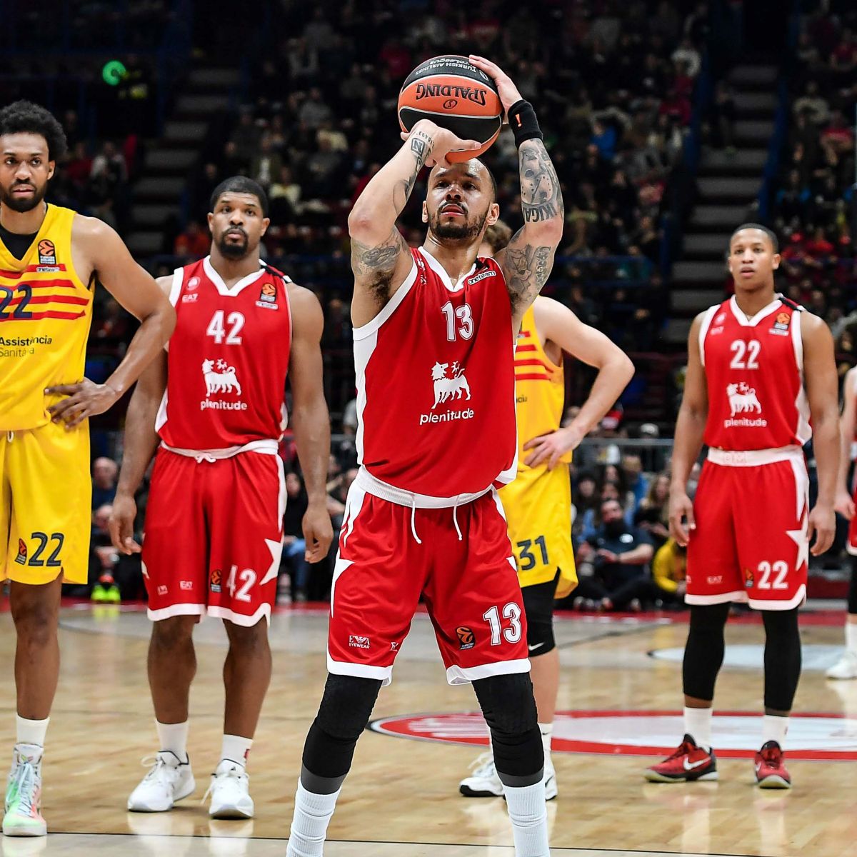In un campo da basket professionale ci sono tre giocatori vestiti di rosso con il logo Plenitude sulla maglia e due giocatori vestiti di giallo, quello con la divisa rossa sta lanciando la palla verso l'alto.