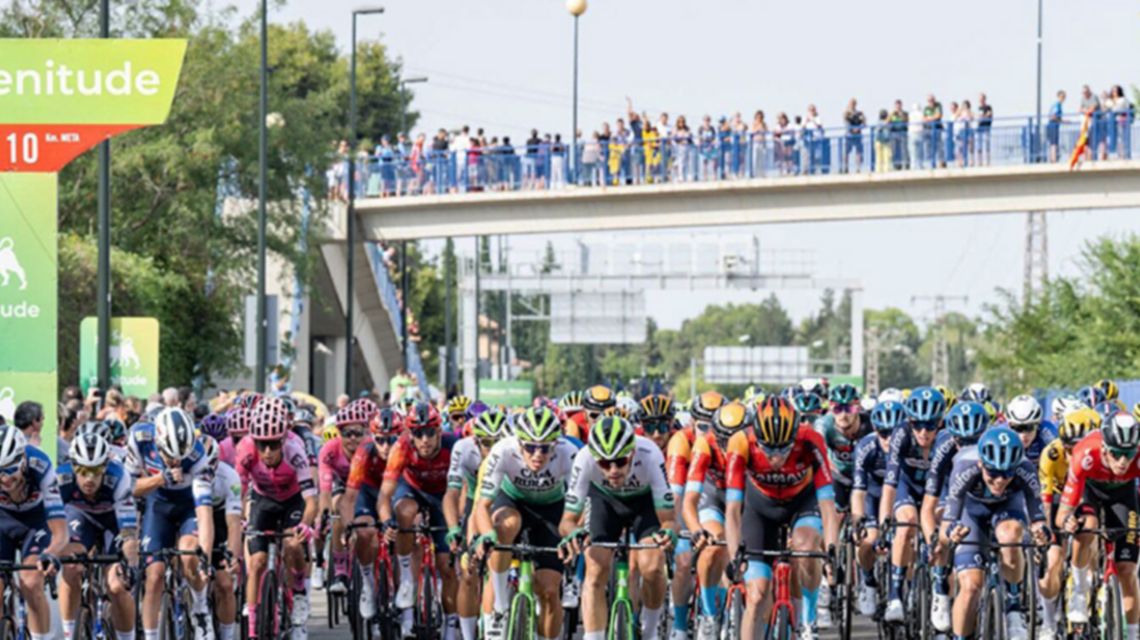 Un gruppo di ciclisti su strada, hanno tutti maglie colorate e pantaloncini da competizione. Sullo sfondo c'è un gruppo di persone che guarda la gara.