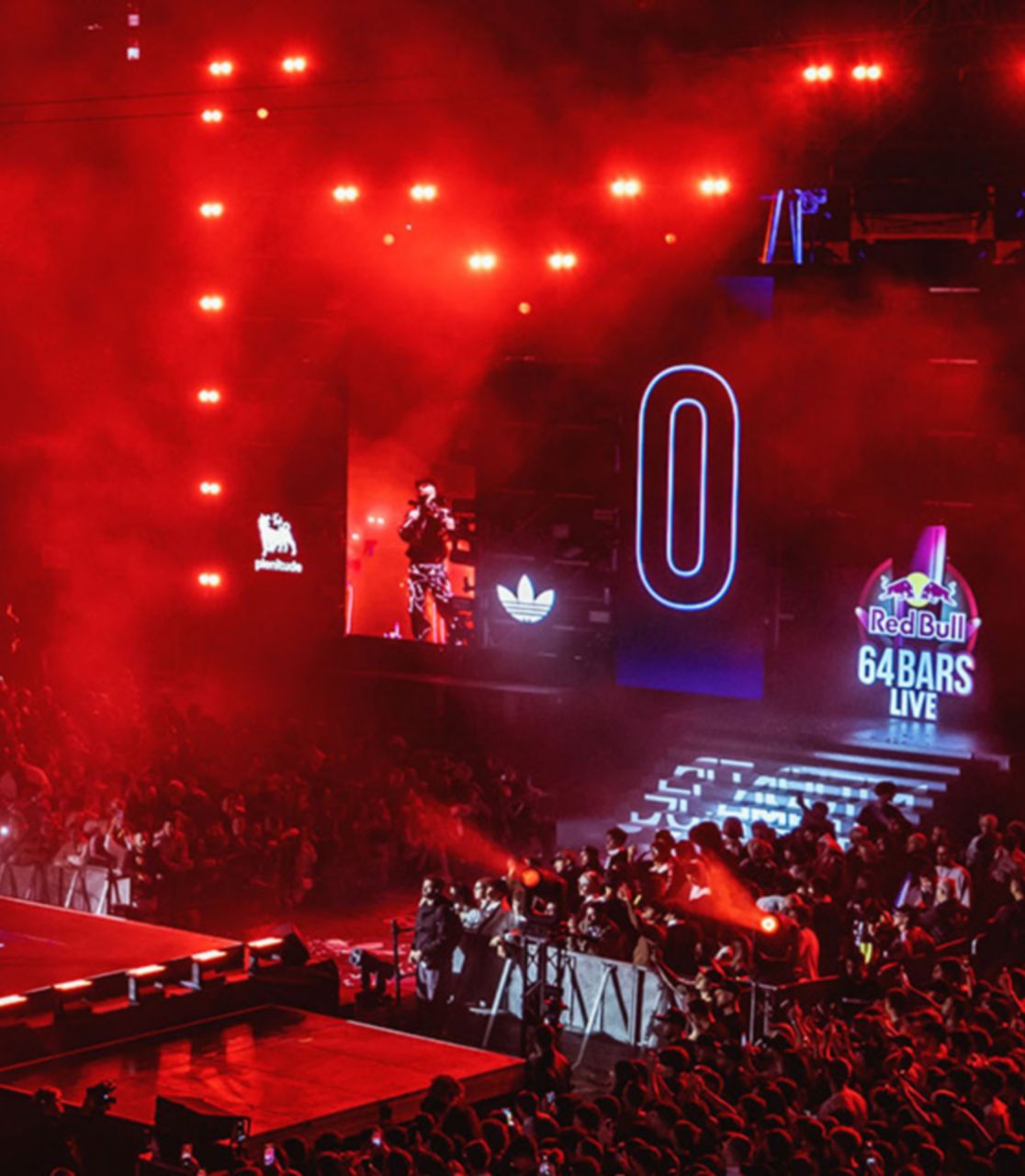 Tra le luci rosse si staglia un palco con delle persone sopra, sull maxi schermo la scritta Red Bull con il logo e 64 Bars live. Intorno al palco c'è una folla di persone, il tutto è visto dall'alto con una ripresa aerea.