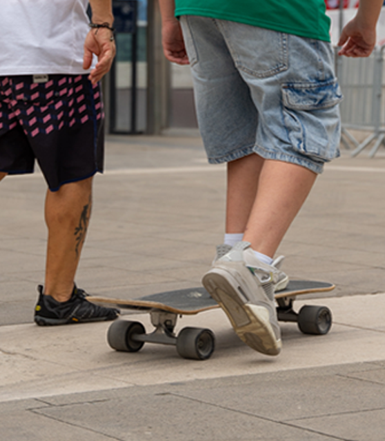 Due ragazzi con pantaloncini e maglietta si trovano in Piazza Duca d'Aosta, all'interno dello skate park. Uno dei due cammina, l'altro è sullo skateboard.