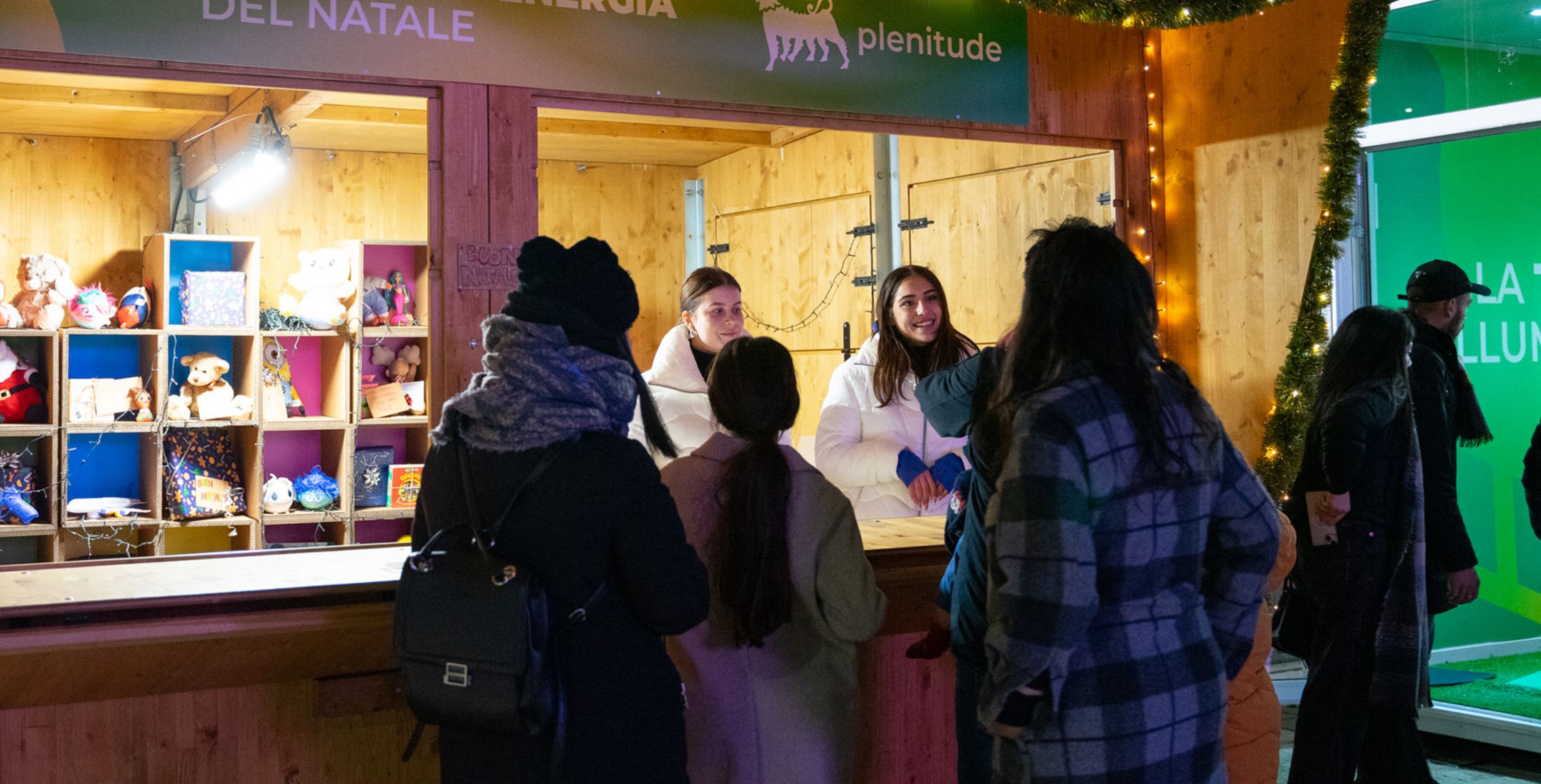 A group of people in winter clothes stand in front of a wooden stand. Inside the stand are two people and shelves with children's games and books. On the stand is the Plenitude logo and the words "Share the energy of Christmas".