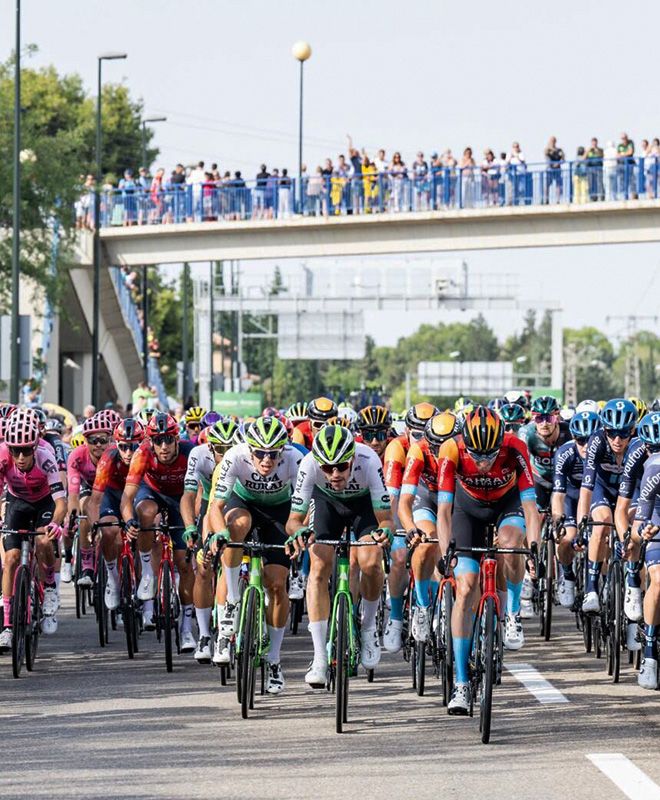 A group of road cyclists, they all have colourful jerseys and racing shorts. In the background is a group of people watching the race.