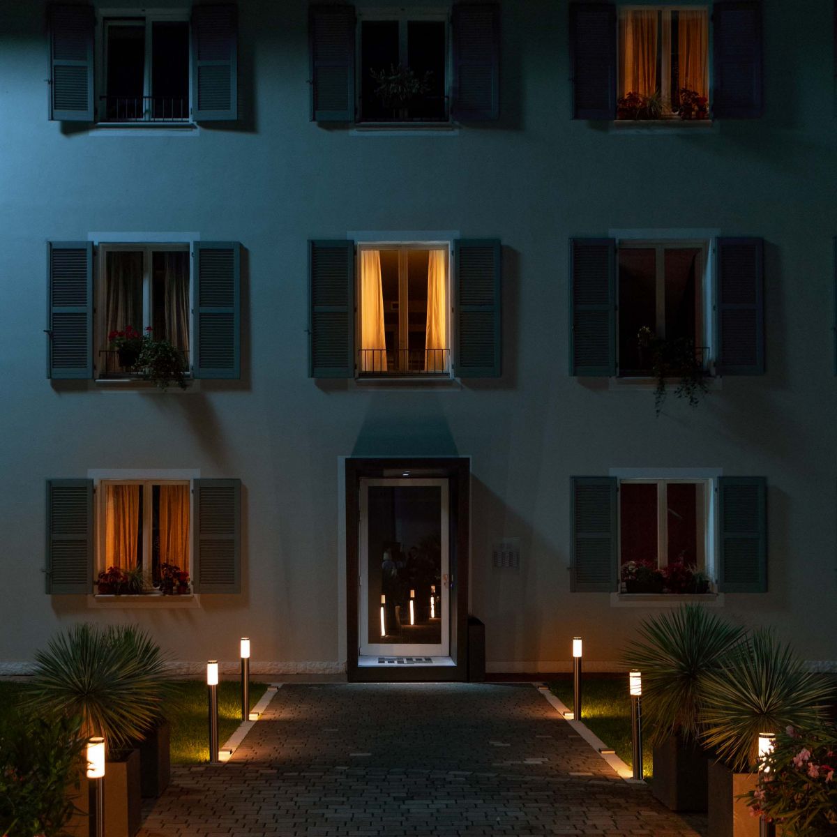Una casa bianca con tre finestre illuminate e le altre al buio. Davanti alla porta d’ingresso c’è un vialetto con delle luci da esterno accese.