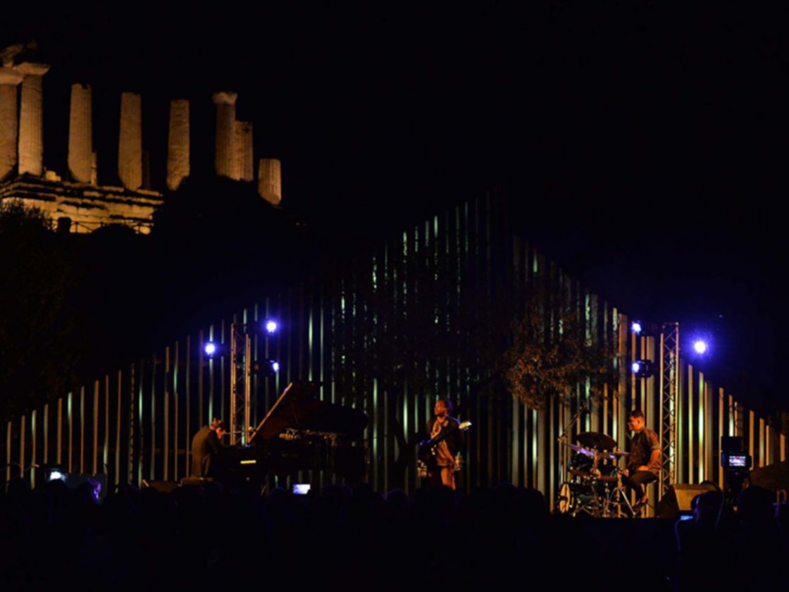 È buio, in uno spazio aperto sullo sfondo ci sono delle rovine antiche, un templio con delle colonne illuminate da fari. In primo piano una band di musicisti su un palco illuminato con un pubblico seduto davanti al buio. 