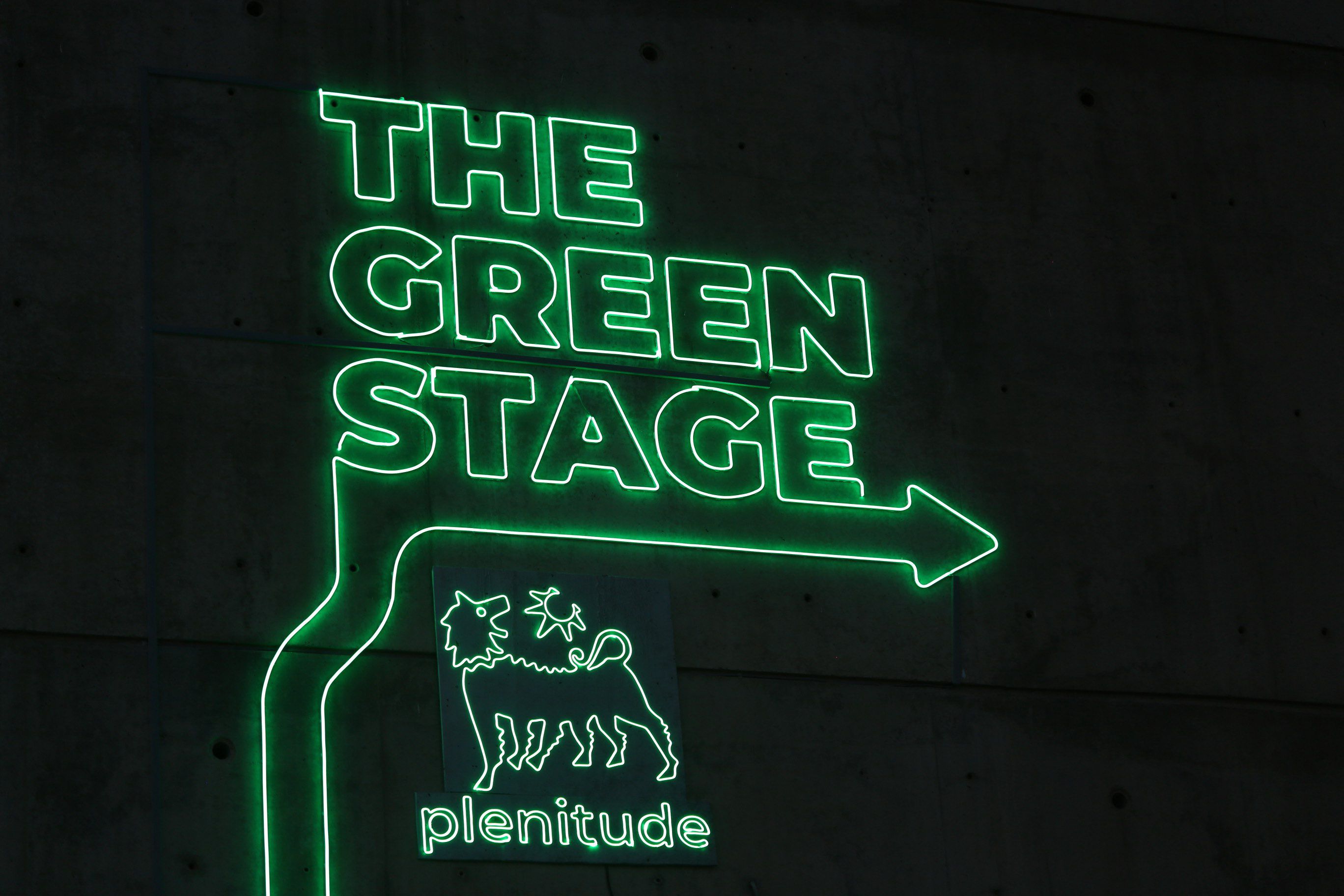 Lettering 'The Green Stage', green, in neon, with an arrow pointing to the right. Below, in green neon, the symbol of Plenitude, a six-legged dog with its face turned towards the sun and the sign 'Plenitude'.