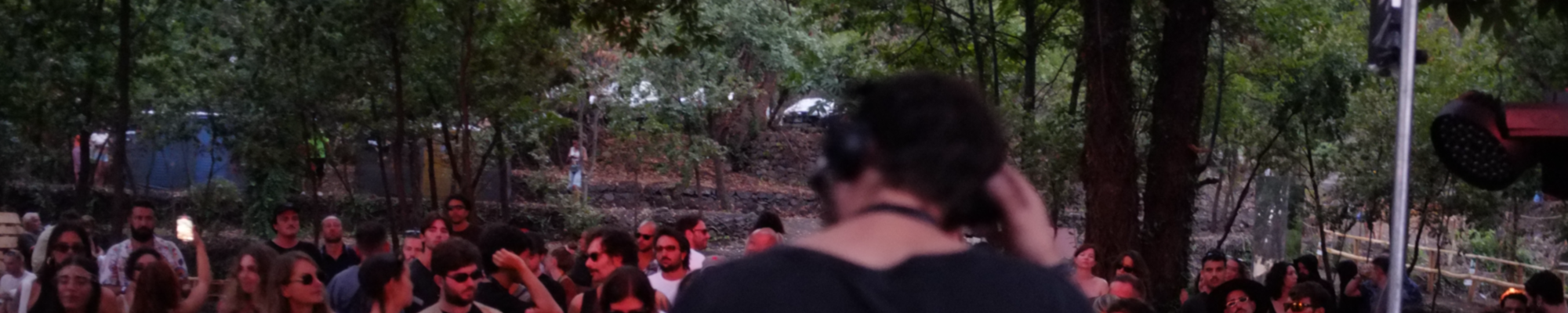 In the foreground, a young man with his back to a pair of black headphones is leaning forward, wearing a dark t-shirt and holding his headphones with his right hand. In the background is a group of boys and girls under a series of green trees.