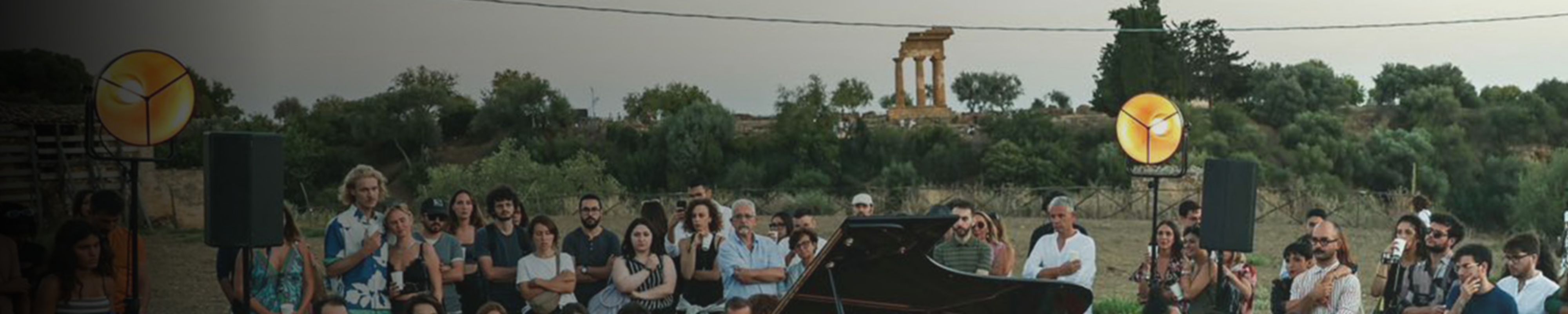 In uno spazio aperto, sullo sfondo di vedono delle rovine antiche, in primo piano una folla seduta è rivolta verso un pianoforte a coda. 