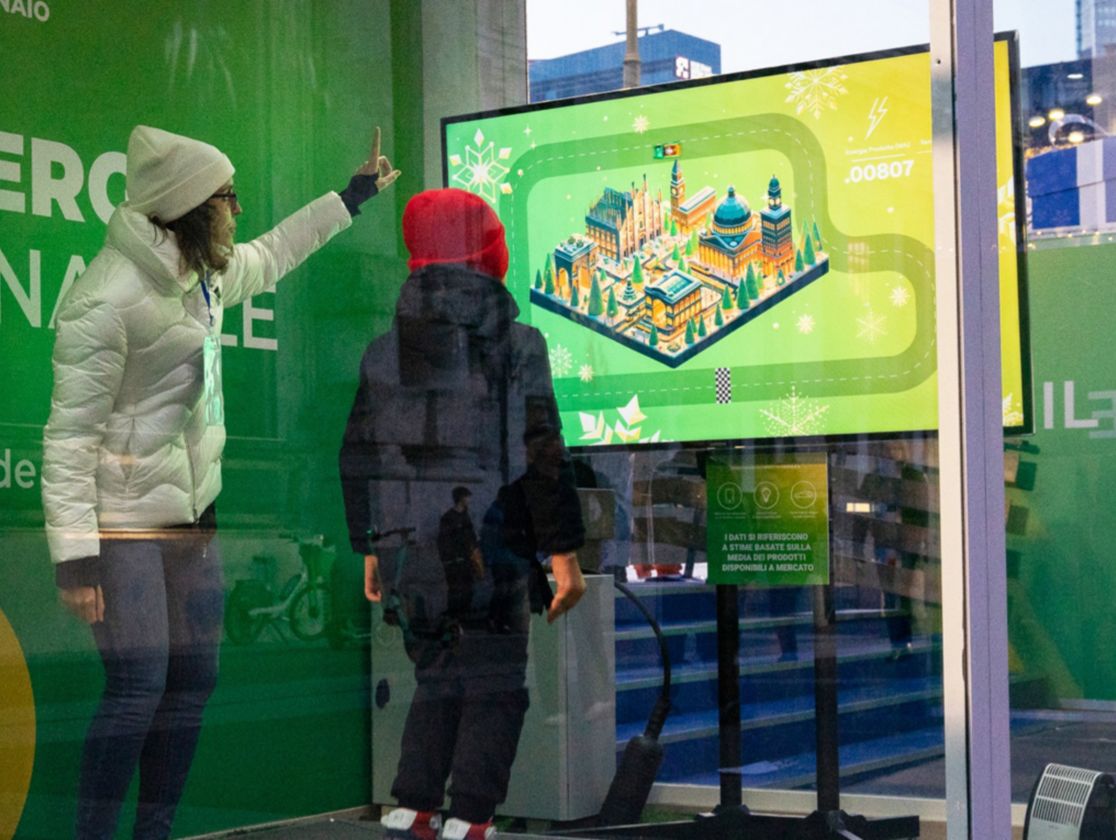 A boy wearing dark winter clothes and a red cap jumps on black platforms while looking at a screen on which a game with a race is projected. Next to him, a girl in blue jeans, a white coat and a white cap points at the screen. Both are inside a narrow space, enclosed by glass windows.