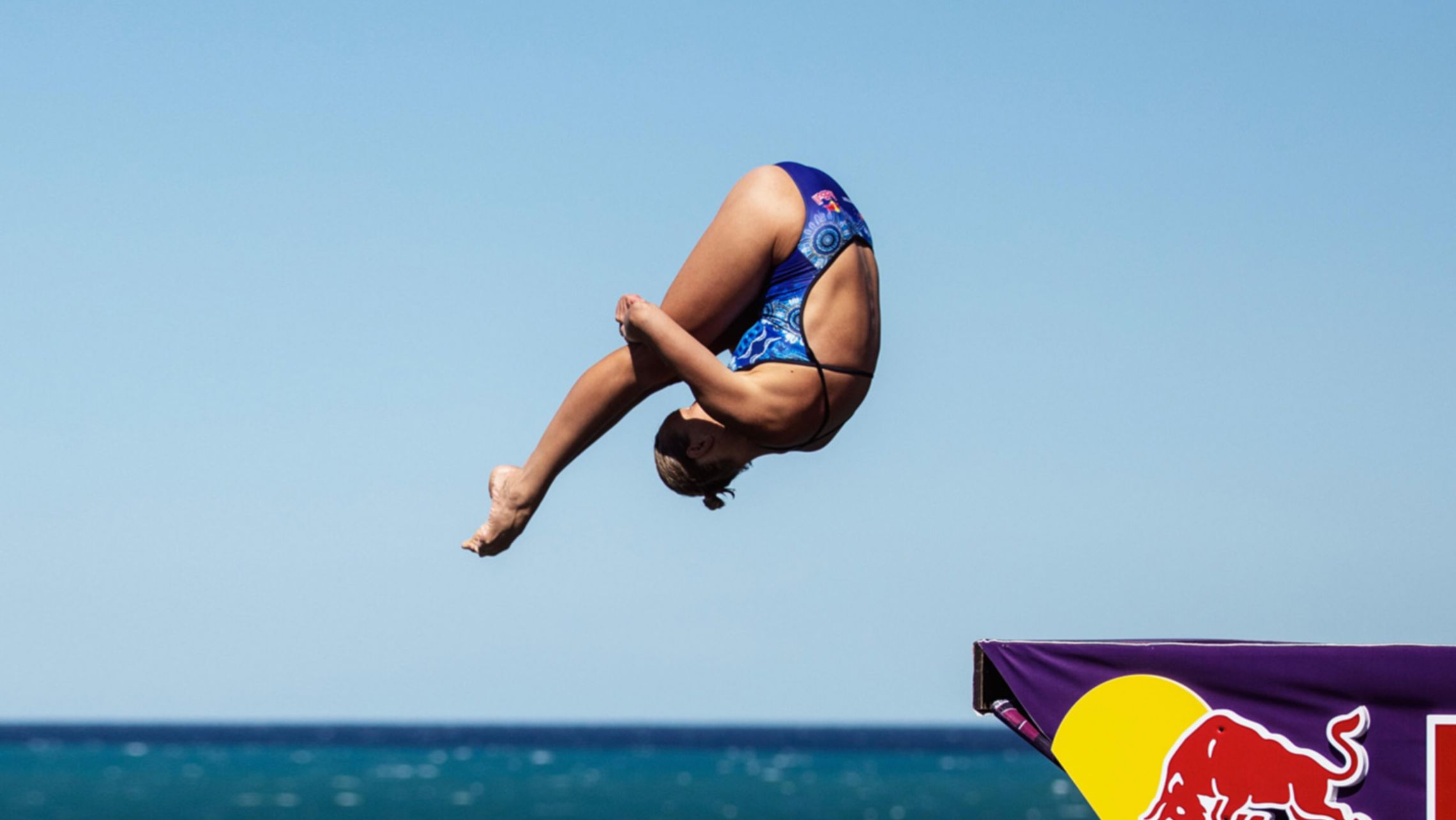 Una ragazza con il costume blu intero è intenta a tuffarsi da un trampolino, su di esso si vede il logo e la scritta di Red Bull.