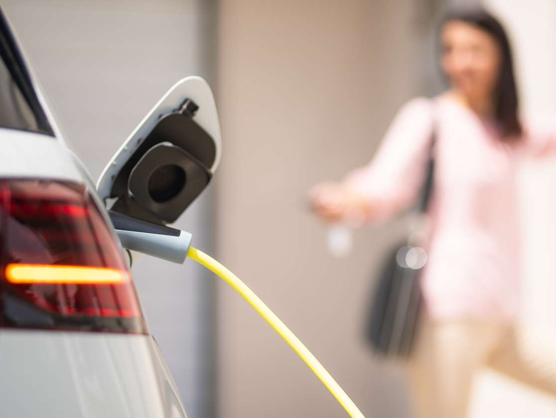 Imagen de un coche conectado a un cargador eléctrico, y una chica detrás