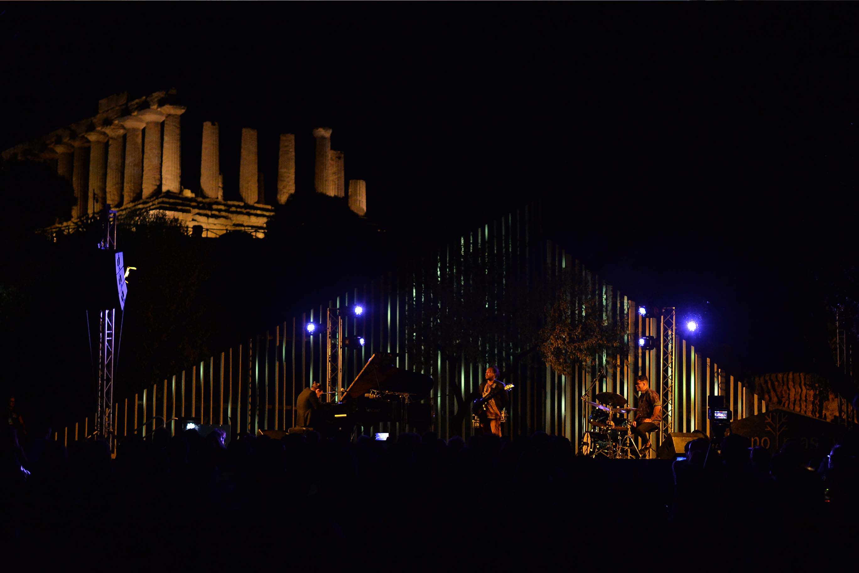 It is dark, in an open space in the background there are ancient ruins, a temple with columns lit by lighthouses. In the foreground a band of musicians on an illuminated stage with a seated audience in front in the dark. 
