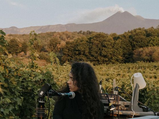 Etna and the nature of the area around the volcano can be seen in the background. In the foreground a girl with dark curly hair, dressed in a long-sleeved black sweater, plays a pianola.
