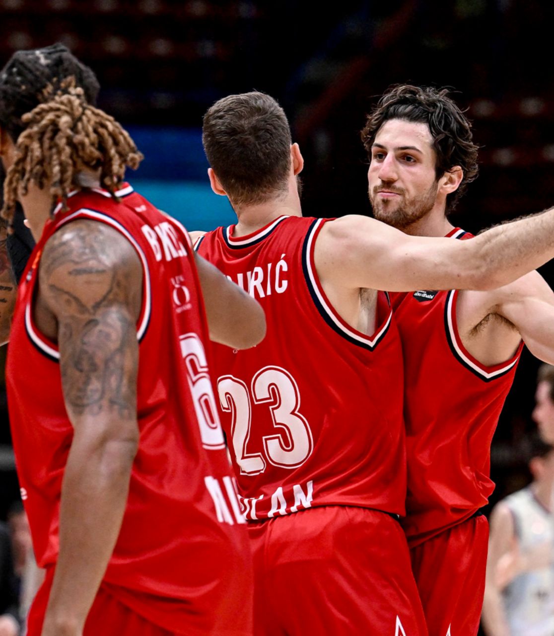 Two basketball players wearing red jerseys with the Plenitude logo are seen in the foreground with their backs turned, facing a third player dressed the same way who looks at them clenching a fist in victory.