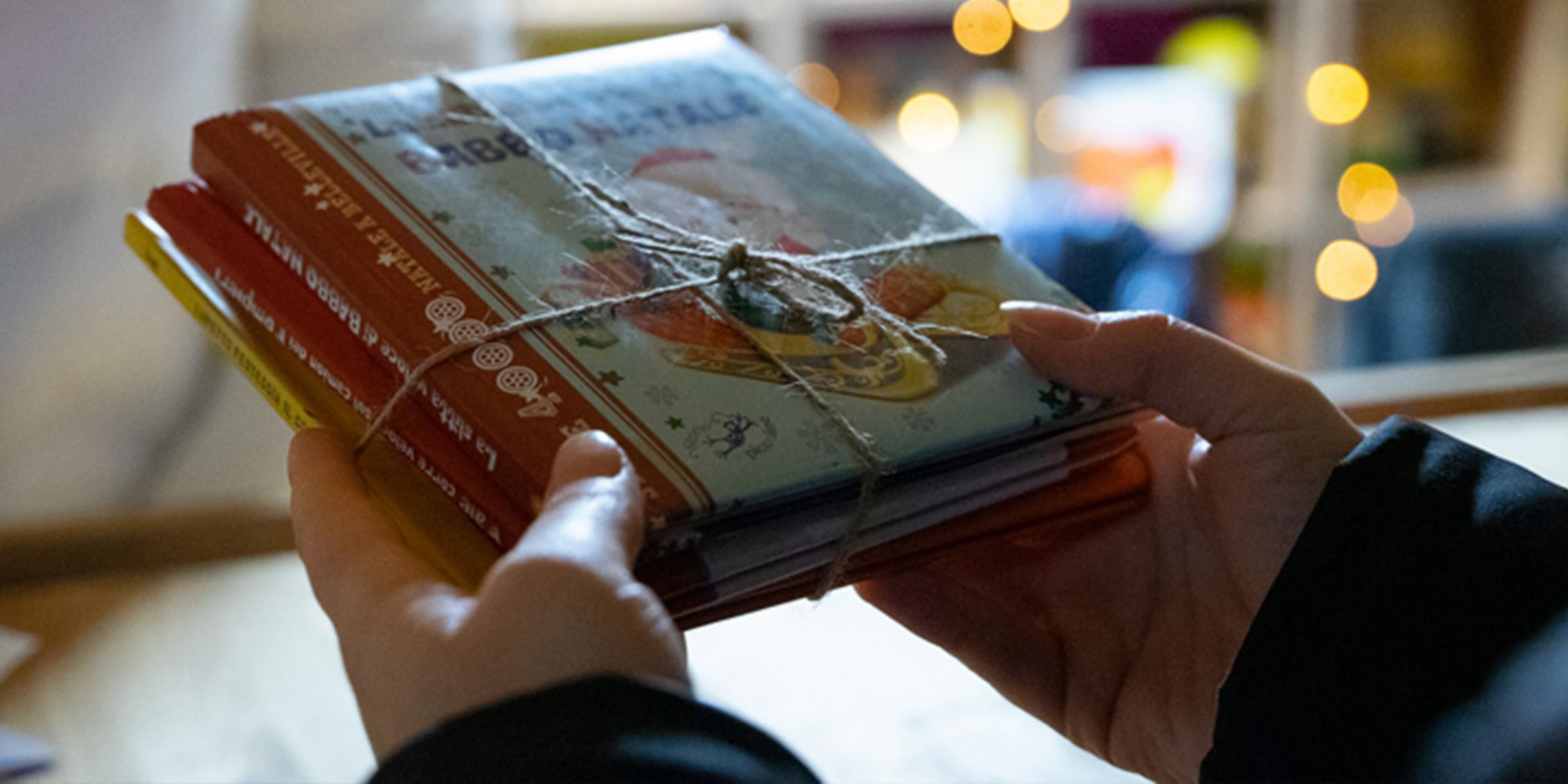 In the background we see an illuminated booth with toys and books inside, a person dressed in white shot from the chin down is beyond the counter. In the foreground a child's hands are holding children's books.
