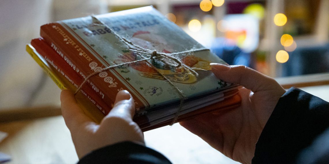 In the background we see an illuminated booth with toys and books inside, a person dressed in white shot from the chin down is beyond the counter. In the foreground a child's hands are holding children's books.