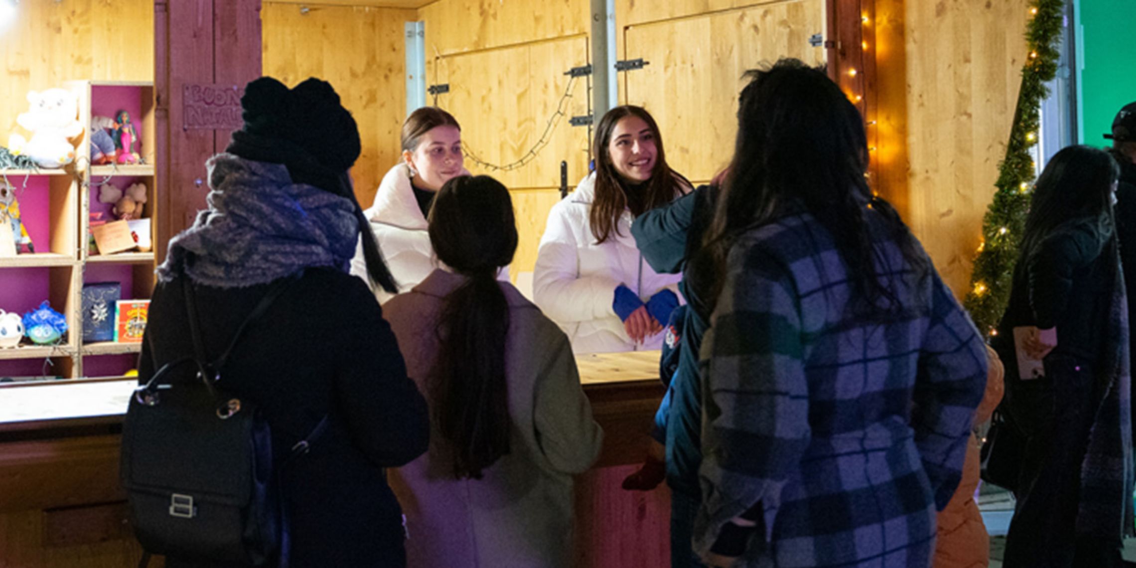 Un gruppo di persone con vestiti invernali si trova davanti ad uno stand di legno, al suo interno ci sono due persone e degli scaffali con giochi e libri per bambini. Sullo stand c'è il logo di Plenitude e la scritta "Condividi l'energia del Natale". 