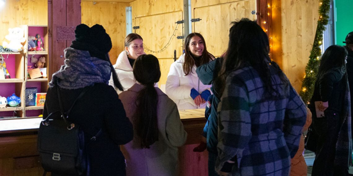 A group of people in winter clothes stand in front of a wooden stand. Inside the stand are two people and shelves with children's games and books. On the stand is the Plenitude logo and the words "Share the energy of Christmas".