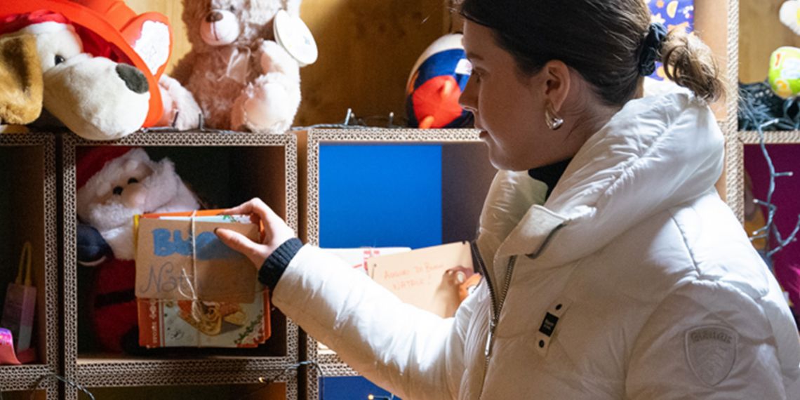A young woman in winter clothes is touching children's books placed on a shelf full of toys. 
