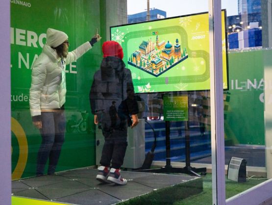 A boy wearing dark winter clothes and a red cap jumps on black platforms while looking at a screen on which a game with a race is projected. Next to him, a girl in blue jeans, a white coat and a white cap points at the screen. Both are inside a narrow space, enclosed by glass windows.