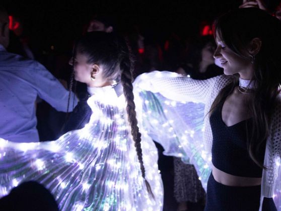 In the middle of a crowd of people are two girls dressed in a black top and trousers, they have wings made from a curtain of clear LED lights.