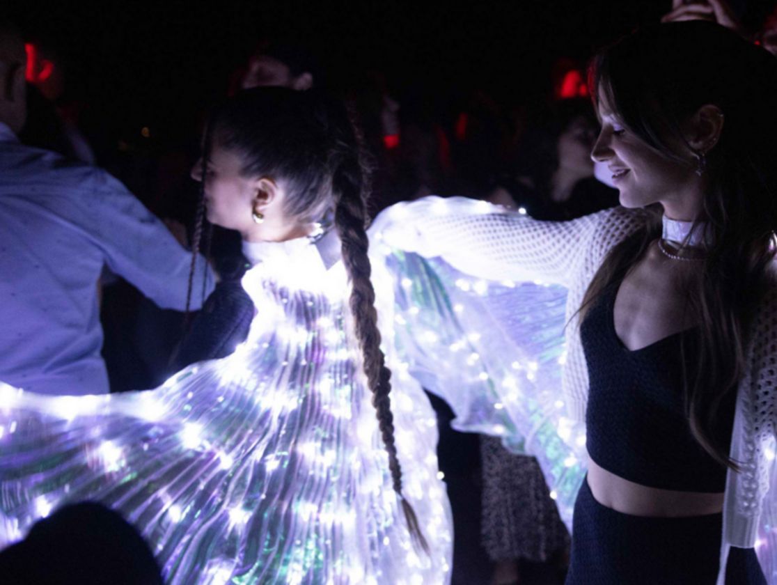 In the middle of a crowd of people are two girls dressed in a black top and trousers, they have wings made from a curtain of clear LED lights.
