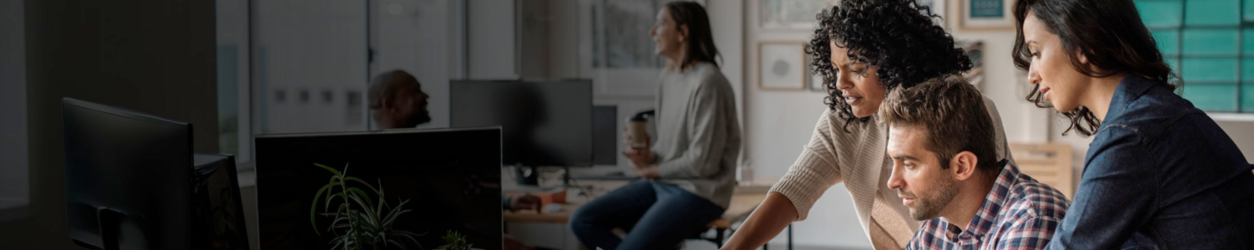In an office there are three people looking at a screen. One is a guy sitting at his desk, his hands on the keyboard, the other two are two women standing behind him, one of them pointing at the screen. In the background a guy sitting at a desk has a coffee in his hand and is talking to someone sitting next to him.