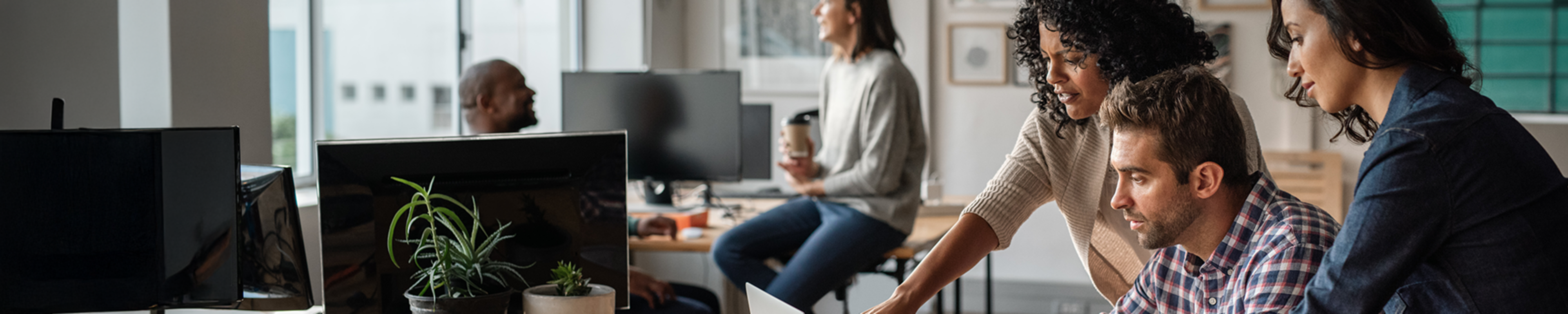 In an office there are three people looking at a screen. One is a guy sitting at his desk, his hands on the keyboard, the other two are two women standing behind him, one of them pointing at the screen. In the background a guy sitting at a desk has a coffee in his hand and is talking to someone sitting next to him.