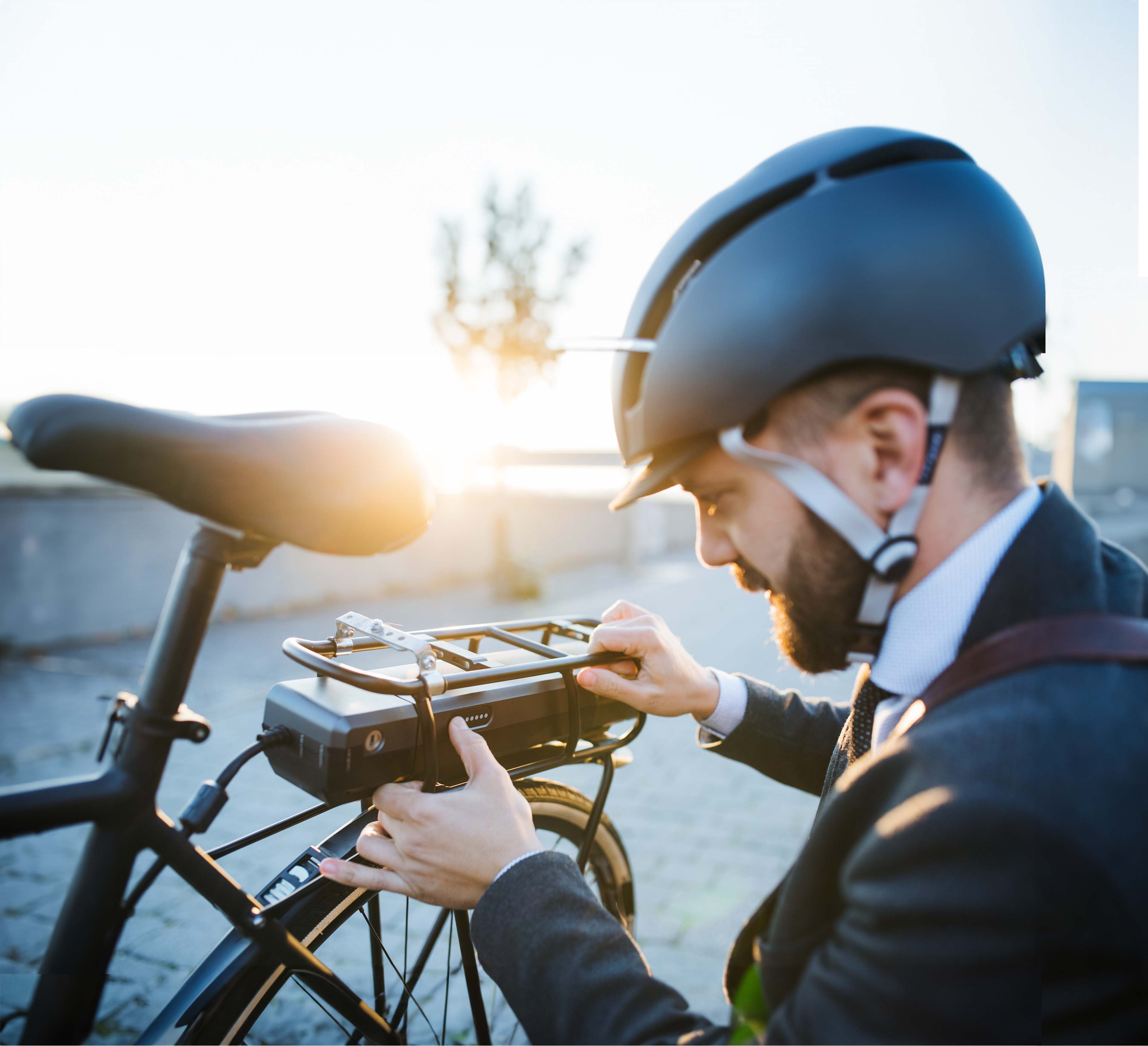 Un uomo vestito in giacca e cravatta, con un casco da bici in testa, è in ginocchio e guarda da vicino il motore di una bici elettrica. 