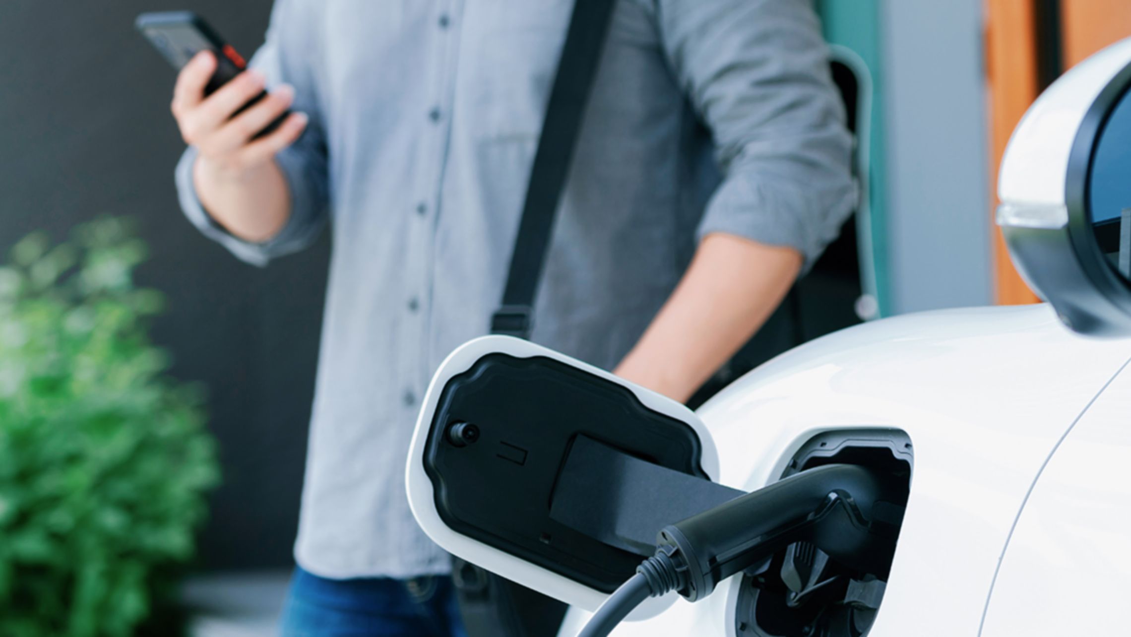 In the foreground a white electric car is charging, in the background a man standing with a phone in his hand.