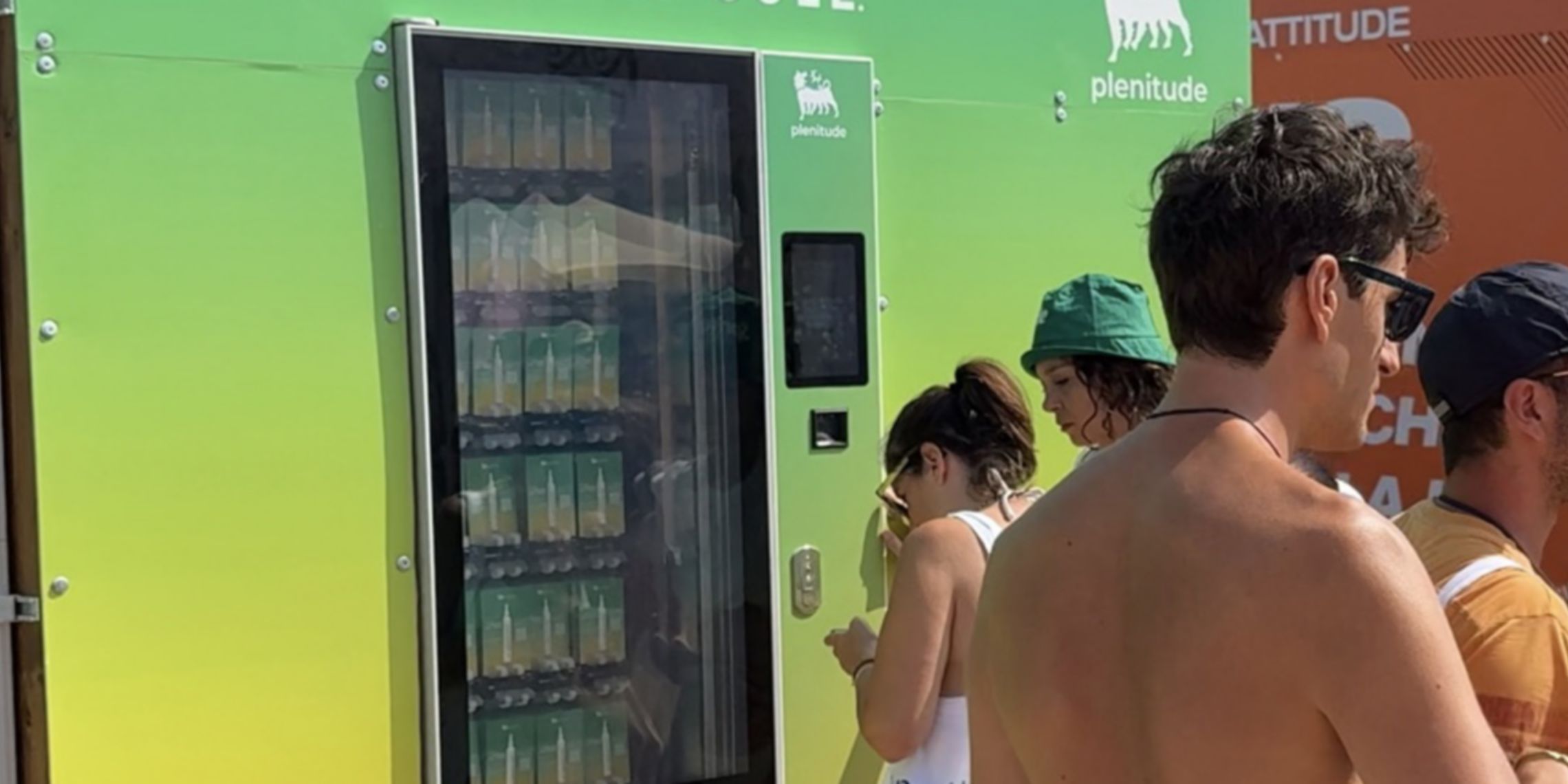 Dei ragazzi in costume da bagno sono davanti ad un distributore automatico verde con il logo e la scritta Plenitude. Al suo interno si intravedono dei campioni di crema solare Plenitude.