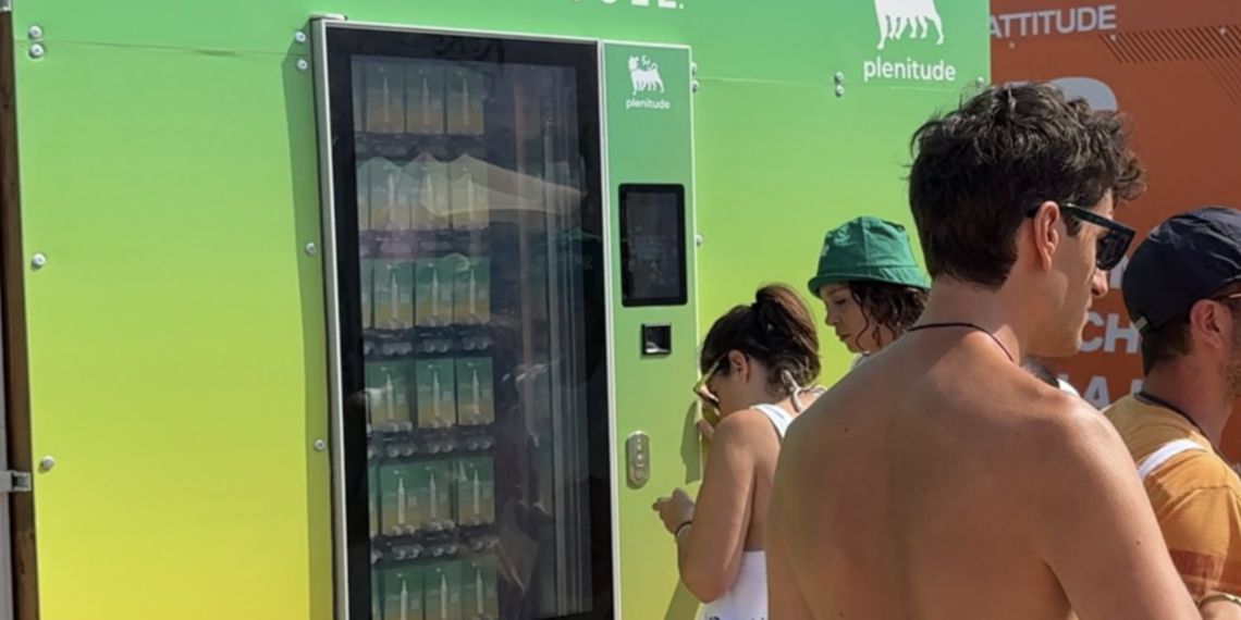 Boys in swimming costumes stand in front of a green vending machine with the Plenitude logo and lettering. Inside, samples of Plenitude sun cream can be glimpsed.