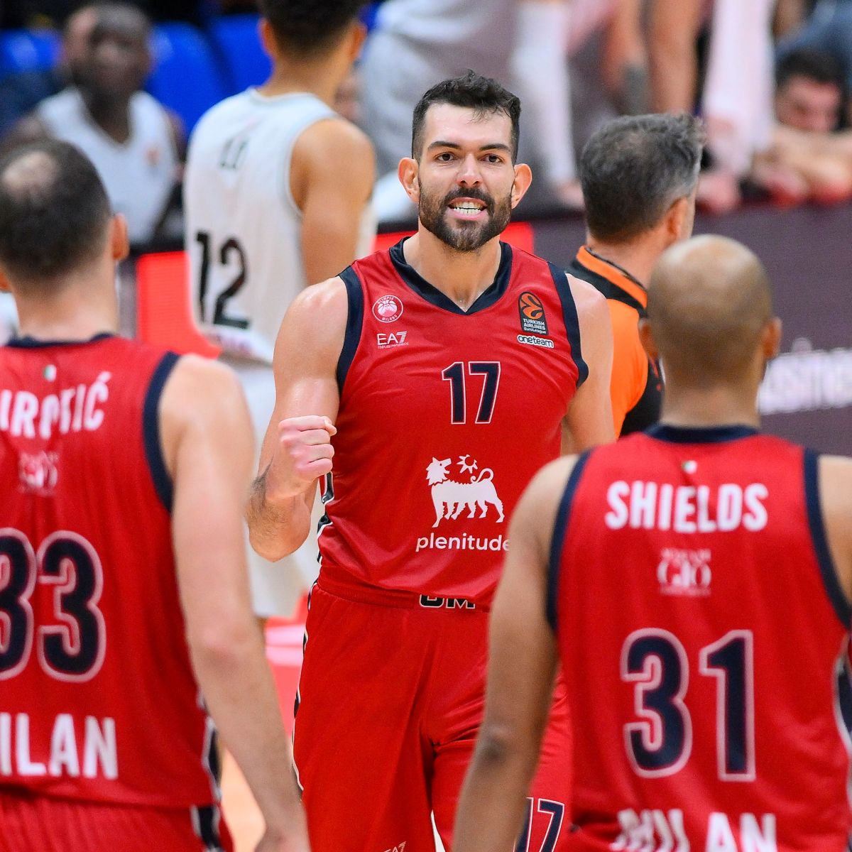 Two basketball players wearing red jerseys with the Plenitude logo are seen in the foreground with their backs turned, facing a third player dressed the same way who looks at them clenching a fist in victory.