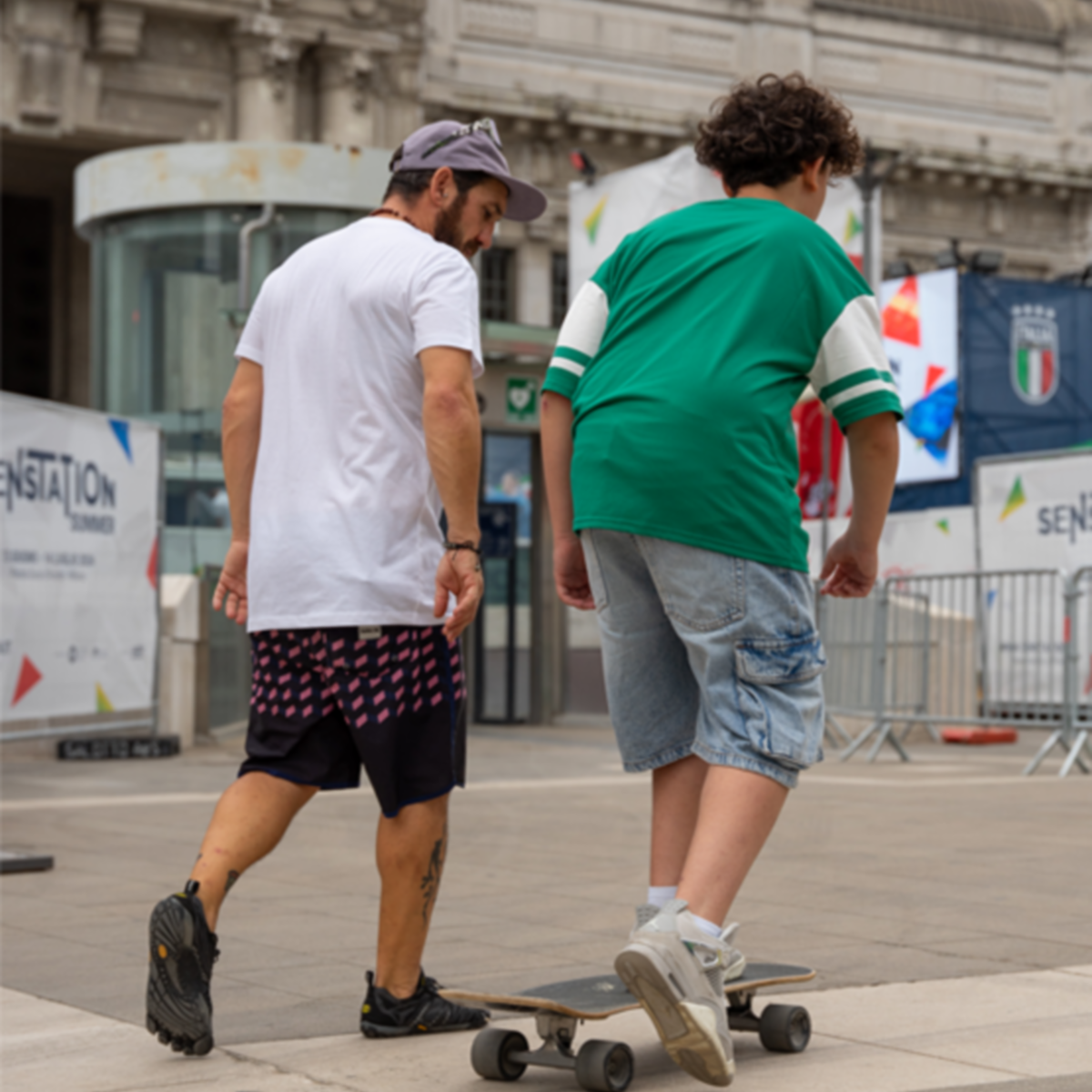 Due ragazzi con pantaloncini e maglietta si trovano in Piazza Duca d'Aosta, all'interno dello skate park. Uno dei due cammina, l'altro è sullo skateboard.