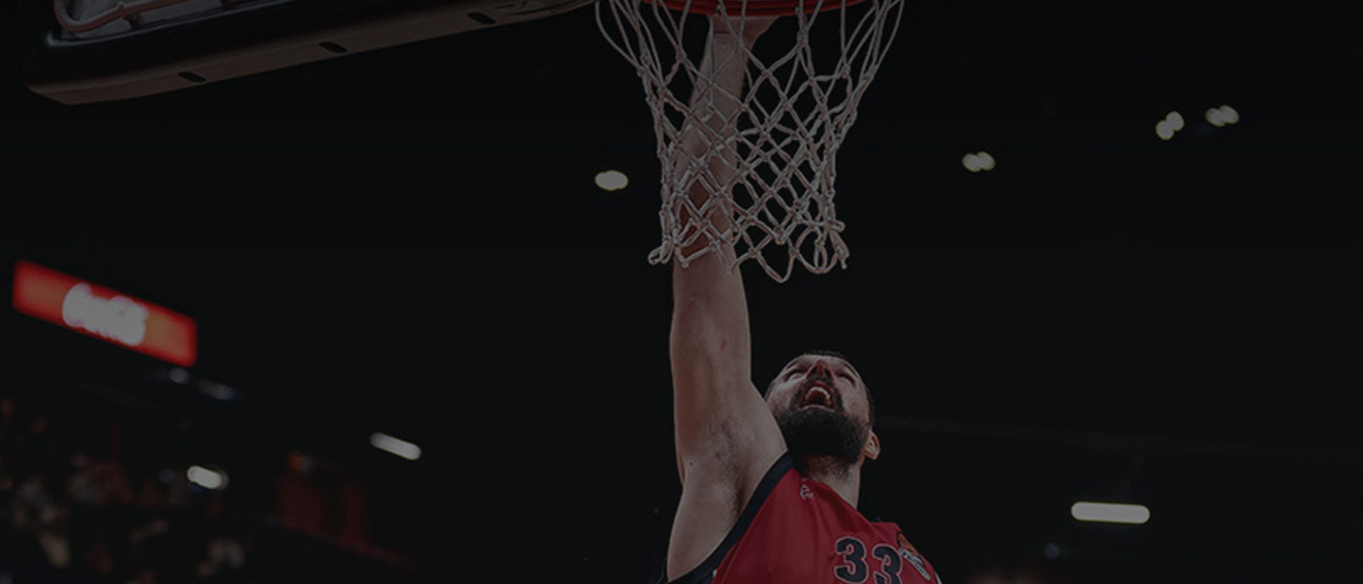 One sees the face of a player with a beard and black hair, dressed in a red basketball vest, scoring a basket with his right arm stretched upwards.