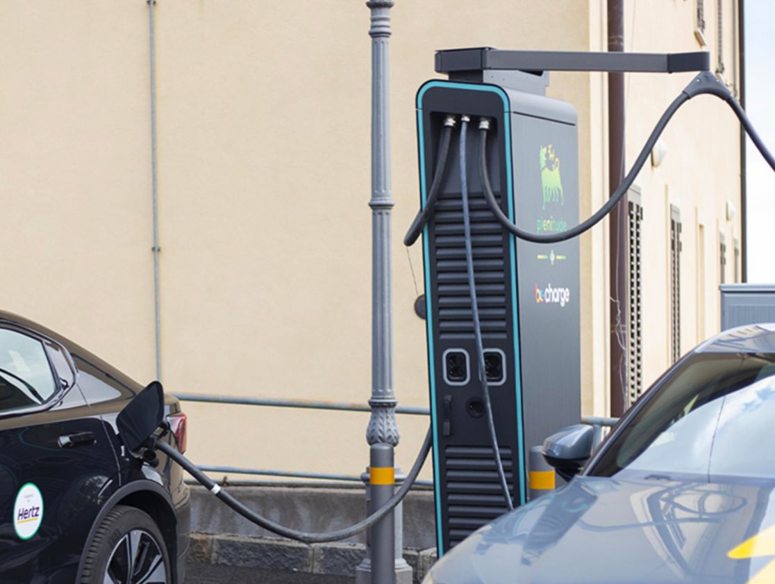 Two electric cars are being recharged at a Plenitude charging station, one is black and one is grey. On the grey one you can see the lightning bolt symbol on the bonnet, on the black one the word Electric and the Plenitude and BeCharge logos.