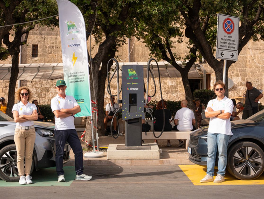 A woman and two men dressed in jeans and a white polo shirt stand in front of a Plenitude charging station and two electric cars are charging. One of the two men is wearing a green Plenitude cap.