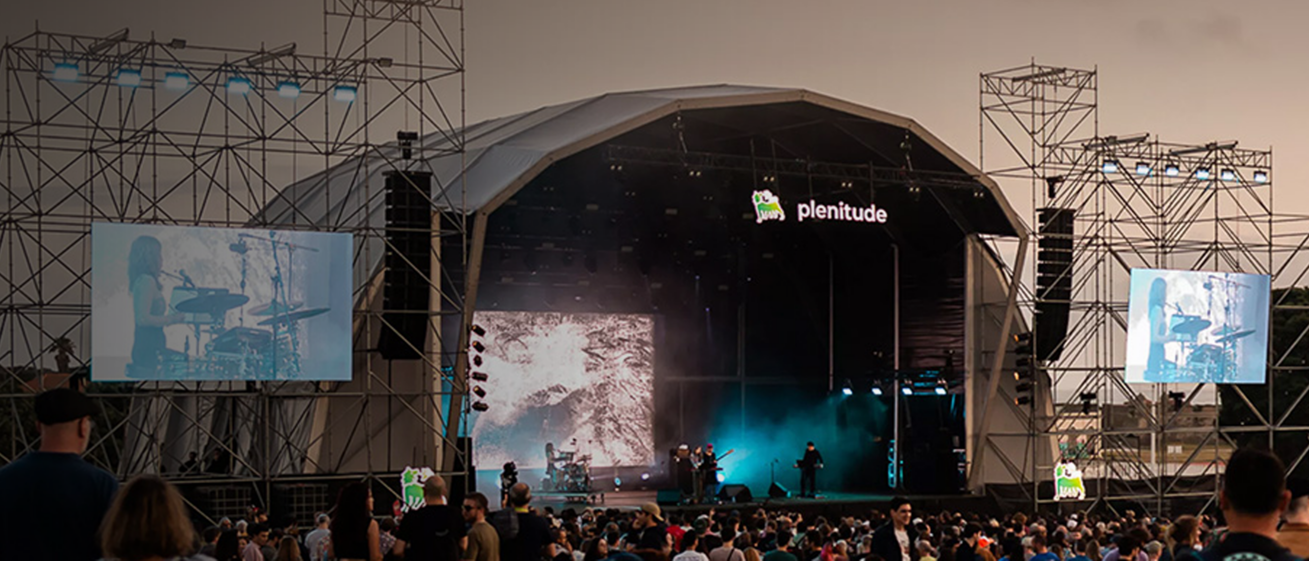 One can see a stage branded with the Plenitude logo and lettering, with a crowd of people in front looking in the direction of the band performing. Behind the band and on the sides of the stage are giant screens showing the show.