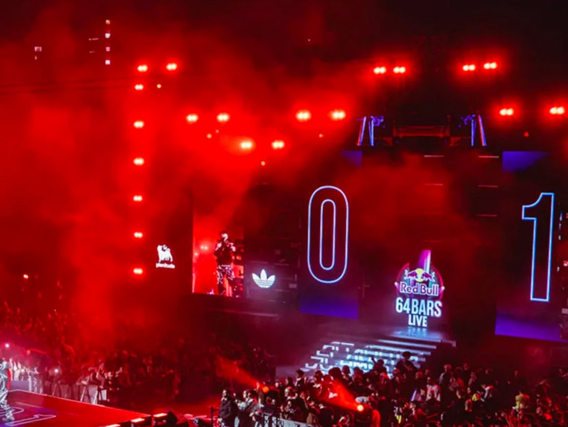 A crowd surrounds a stage where there is a person holding a microphone, the screen at the back reads RedBull 64 bars live.