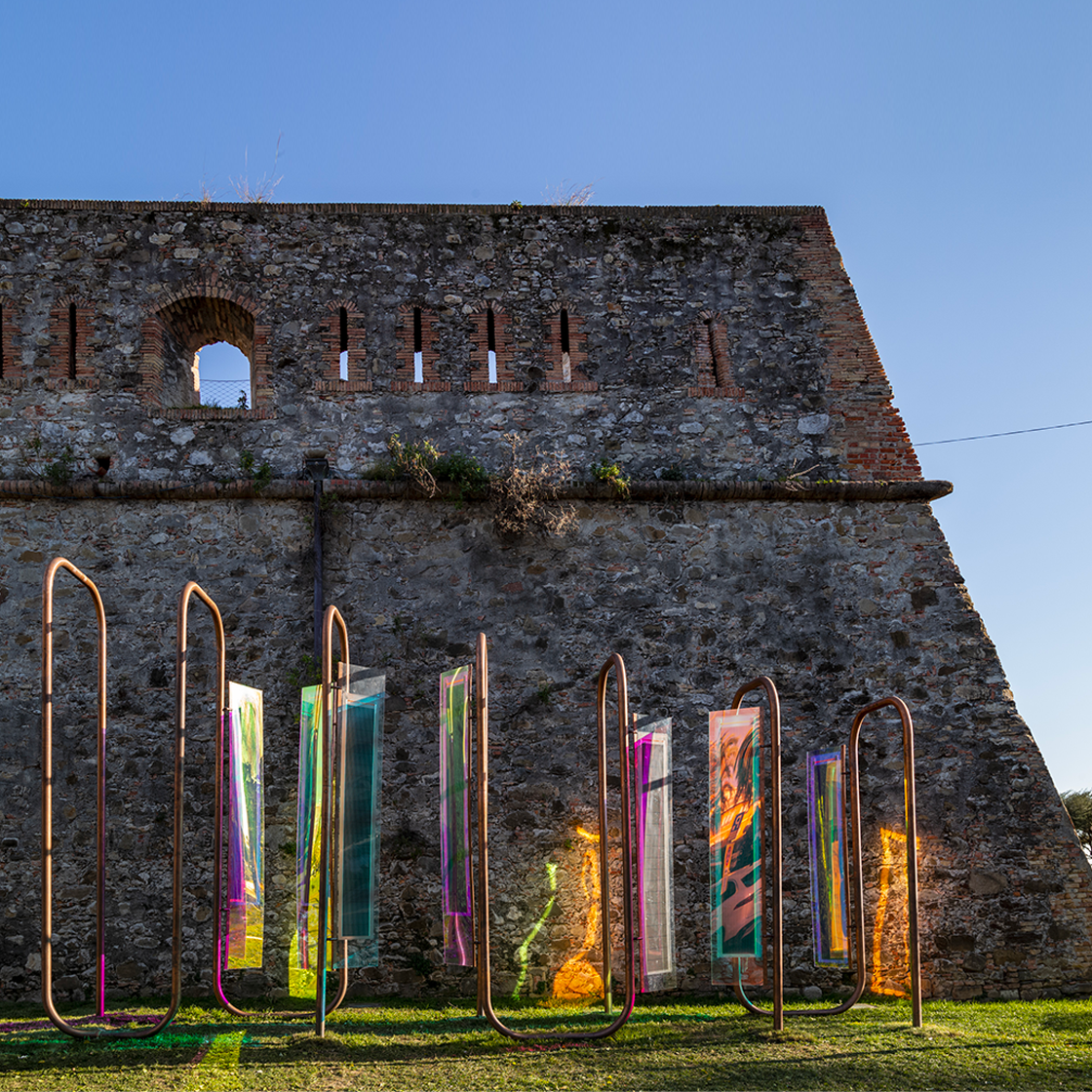 Picture of a coloured metal installation in the foreground, an ancient wall in the background.