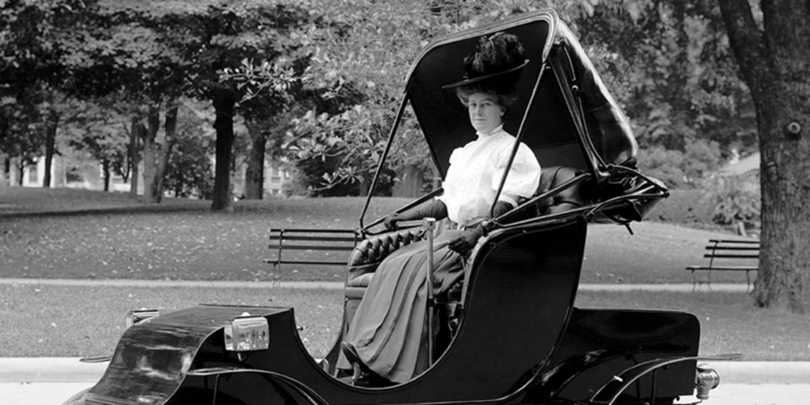 Una signora con abiti ottocenteschi è seduta su una carrozza in una foto in bianco e nero.